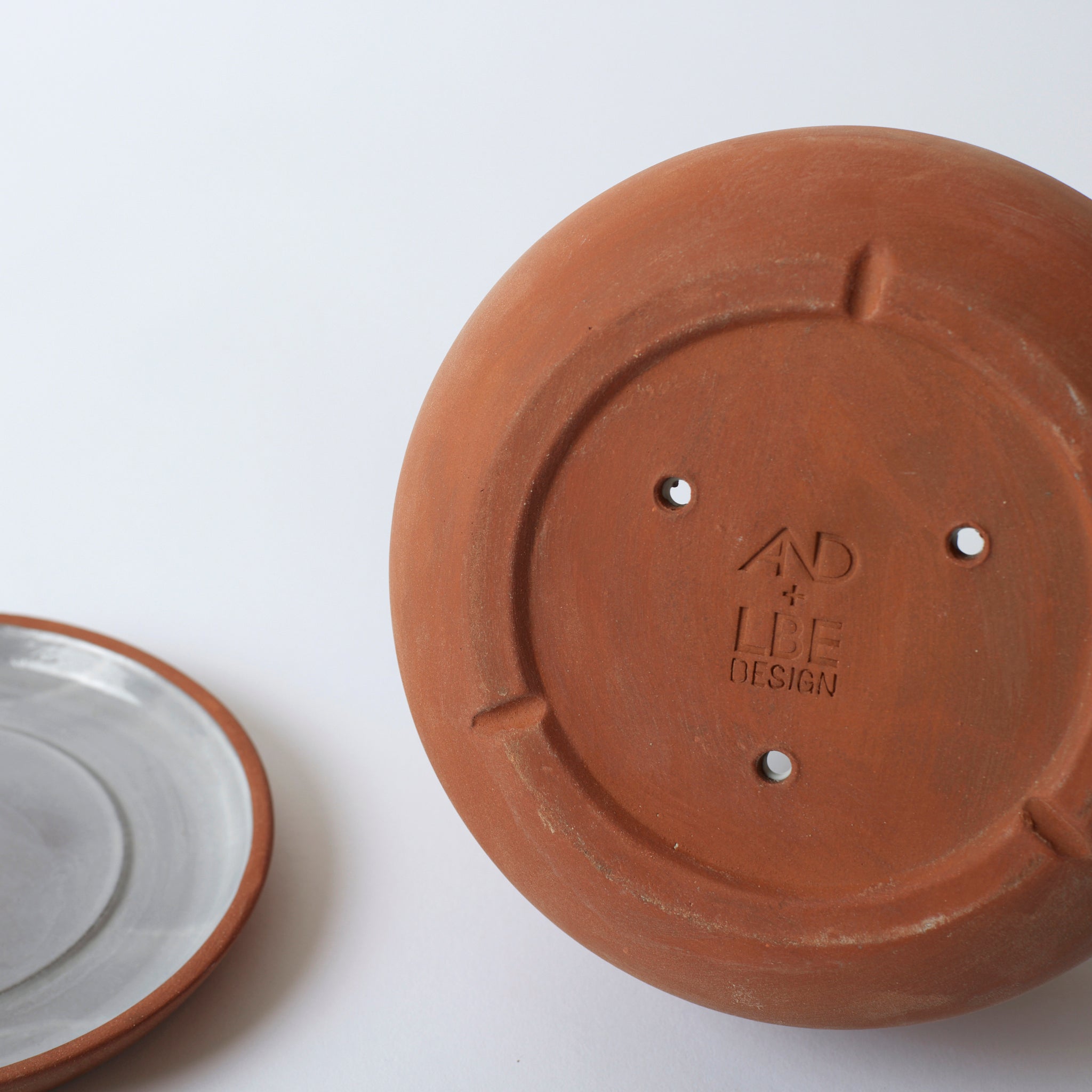 Close up image of the bottom of the terra cotta Rancho Planter. It features drainage holes and a stamped logo reading "AND + LBE Design".